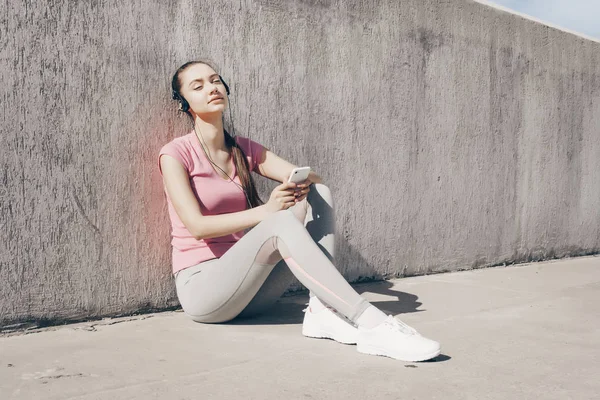 Втомлена струнка дівчина в рожевій футболці сидить на землі на сонці і слухає музику на навушниках, відпочиваючи після бігу — стокове фото