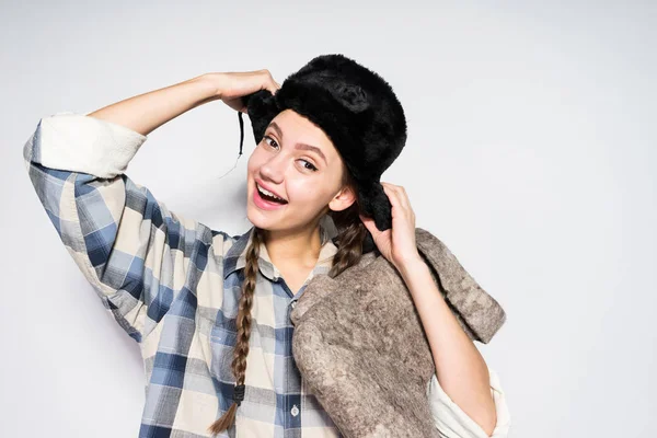 Una divertida chica rusa con coletas sonríe y sostiene botas de fieltro gris cálido, un sombrero de piel negro en su cabeza — Foto de Stock