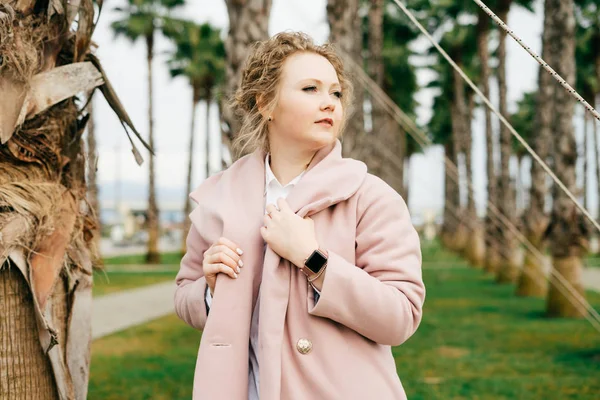 Elegant beautiful woman in a stylish pink coat enjoying a warm spring in a park next to green palms — Stock Photo, Image