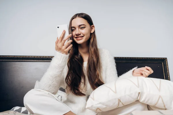 Милая девушка с длинными волосами в белой пижаме сидит на кровати рано утром, улыбаясь и глядя на свой смартфон — стоковое фото