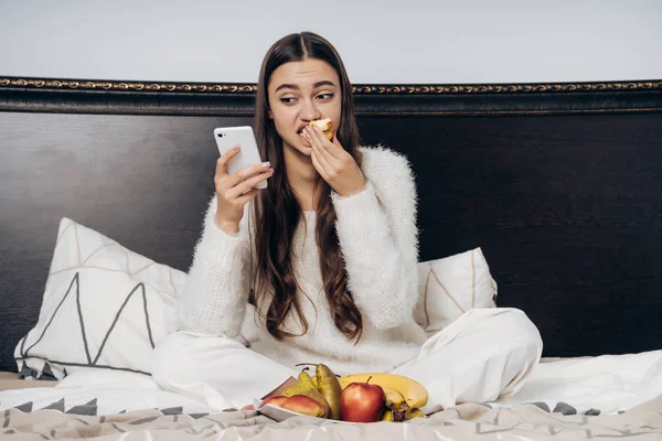 Joven en pijama blanco se sienta en la cama, come fruta útil y mira en su teléfono inteligente — Foto de Stock