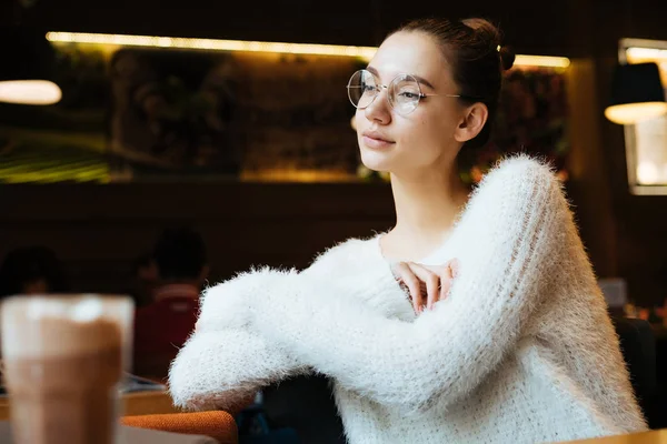 Carino giovane studentessa in un maglione bianco e occhiali a riposo in un caffè, guardando fuori dalla finestra — Foto Stock