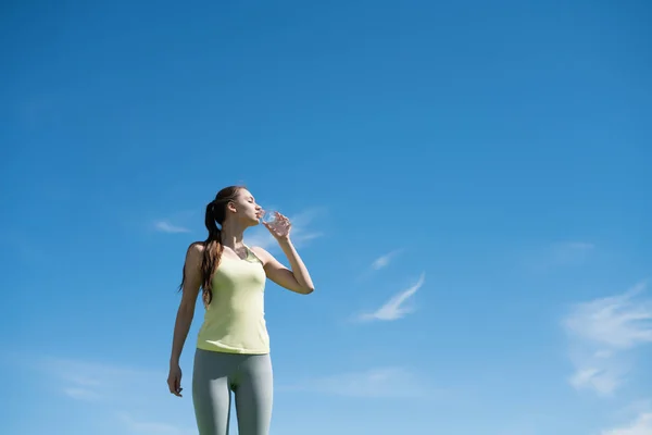 Krásná štíhlá holka pije čistou vodu po těžké cvičení, pod modrou oblohou — Stock fotografie