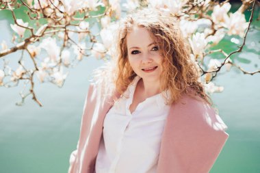 bahar Isı kıvırcık zarif kadının bir gölet, yanındaki bir parkta poz pembe bir ceket