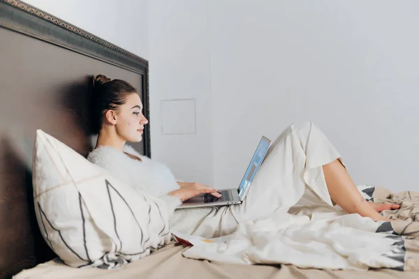 Schattig jong meisje in witte pyjama zitten in bed laat op de avond, kijken naar Tv-serie op laptop — Stockfoto