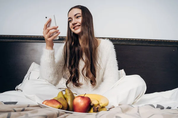 Nettes schönes Mädchen mit langen Haaren sitzt auf dem Bett, schaut in das Smartphone und isst gesundes Obst — Stockfoto