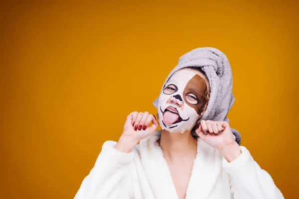 A funny young girl in a white terry dressing gown and with a towel on her head looks after herself, on her face a moisturizing mask with a dog's face — Stock Photo, Image