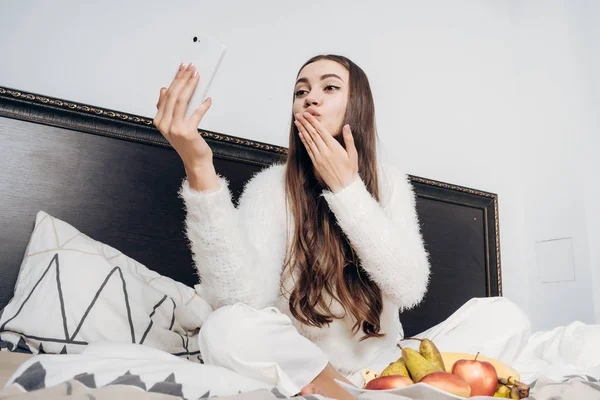 Hermosa chica de pelo largo en pijama blanco se despertó temprano en la mañana, se sienta en la cama, hace selfie en su teléfono inteligente — Foto de Stock