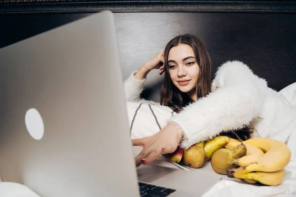 Schattig jong meisje ligt op bed kijken naar een interessante Tv-show op haar laptop en het eten van fruit — Stockfoto