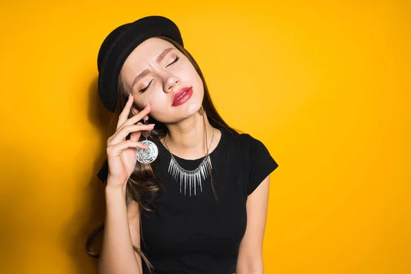 Uma menina bonita luxuosa em um chapéu preto da forma, posando em um fundo amarelo, no pescoço um colar caro — Fotografia de Stock