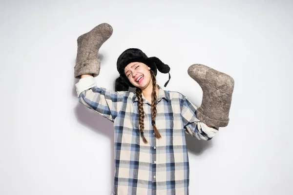 Riéndose chica rusa con coletas en un sombrero de piel caliente se regocija en el invierno, mantiene las botas de fieltro gris en sus manos — Foto de Stock