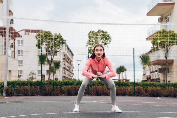 Koppig sterke meisje in een roze jas maakt oefeningen op het sportveld, wil een mooi figuur — Stockfoto