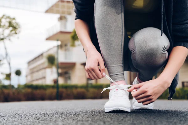 Ein schlankes Mädchen in grauer Sporthose schnürt ihre Schnürsenkel an Turnschuhen, bereitet sich auf einen Lauf vor — Stockfoto