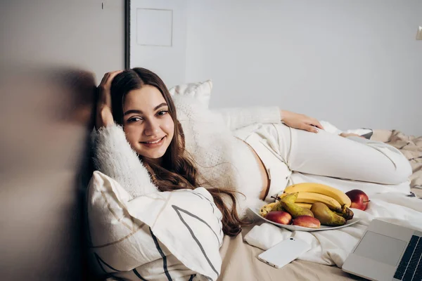 Hermosa chica somnolienta en pijama blanco descansando en la cama después de un duro día de trabajo, sonriendo — Foto de Stock