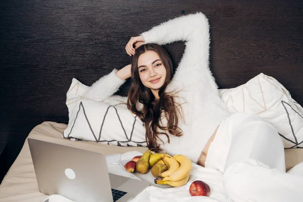 En söt leende flicka med långa tjocka hår ligger i säng tidigt på morgonen, frukost med användbara frukter och ser på en film på en bärbar dator — Stockfoto