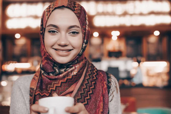 A charming Muslim girl with a beautiful headscarf ate in a cozy restaurant, drinks a fragrant coffee and smiles — Stock Photo, Image