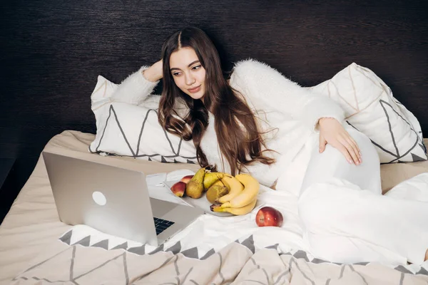 Una chica cansada con el pelo largo yace en la cama, descansando después de un duro día de trabajo y viendo una película divertida en su computadora portátil — Foto de Stock