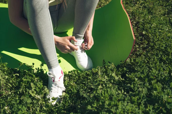 Una chica delgada en pantalones deportivos grises se sienta en una alfombra para el yoga, en un parque sobre hierba verde, lleva un estilo de vida saludable — Foto de Stock