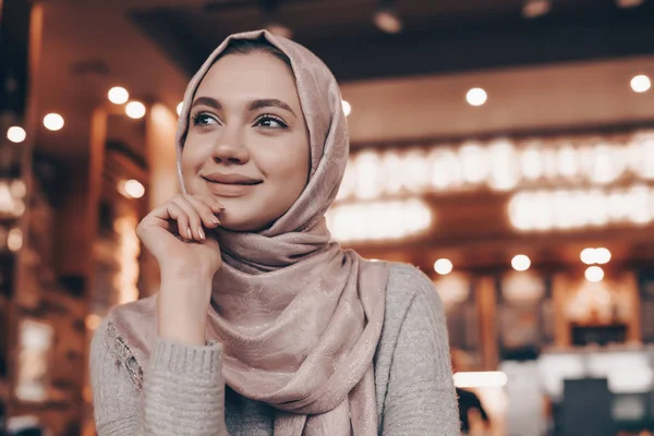Charming young Arab girl in hijab sits in a restaurant, dreams about something and smiles — Stock Photo, Image