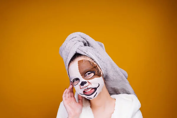 Divertida joven con una bata de felpa blanca, con una toalla en la cabeza sonriendo, en su cara una máscara hidratante con la cara de un perro — Foto de Stock