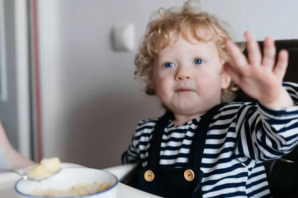 Молодой голубоглазый кудрявый ребенок мальчик ест полезную кашу, семейный завтрак — стоковое фото