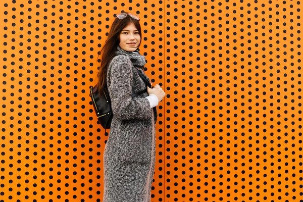 Élégante fille aux cheveux foncés en manteau de mode gris posant près d'un mur orange inhabituel — Photo