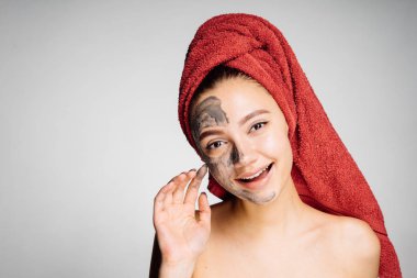 Başında kırmızı bir havlu ile güzel bir kız için yarım yüz yararlı kil maskesi yüzü, cildin uygulayıp sonra görünüyor