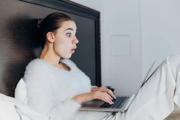 Молода здивована дівчина виглядає цікавим фільмом на своєму ноутбуці, сидить у ліжку під білою ковдрою — стокове фото