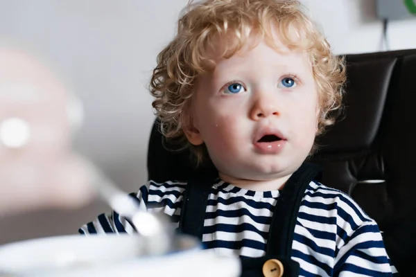 Милый маленький голубоглазый мальчик с светлыми волосами сидит на кухне и смотрит где-то, семейный завтрак — стоковое фото