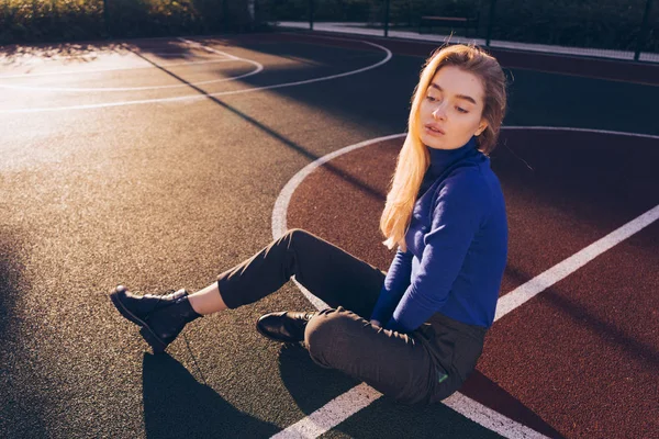 Confiante loira menina modelo em um suéter azul posando em um campo de esportes nos raios do sol da manhã — Fotografia de Stock