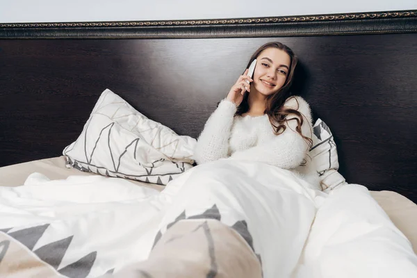 迷人的年轻女孩躺在床下一条白色的毯子, 通话和微笑 — 图库照片