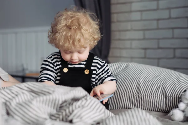 Malý kudrnatý chlapeček v roztomilé oblečení, sedí na posteli a hraje — Stock fotografie