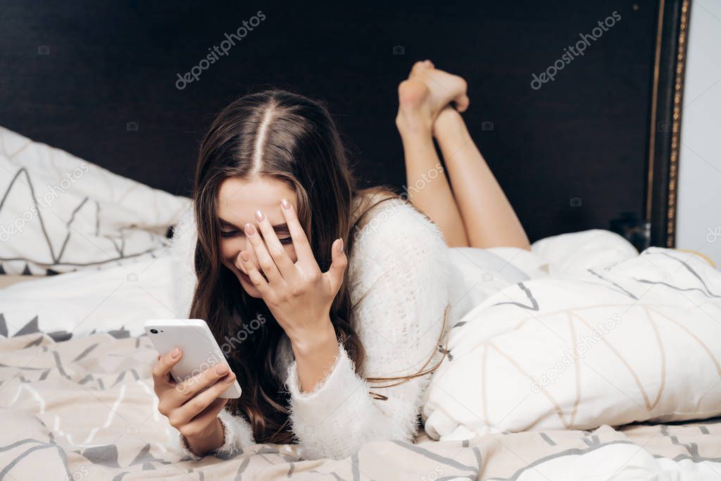 sleepy young girl in white pajamas lies on a bed in the evening, holds a smartphone