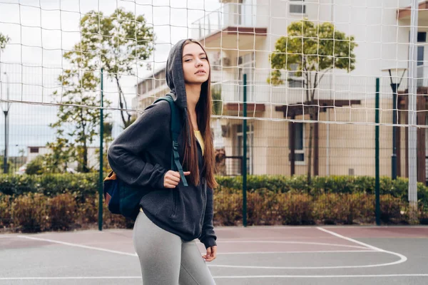 Menina esbelta confiante em uma camisola preta com um capuz segura uma mochila, descansando após o treinamento no campo esportivo — Fotografia de Stock