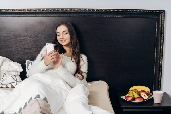 Encantadora joven se encuentra en la cama debajo de una manta blanca, descansando, viendo vídeo en su teléfono inteligente — Foto de Stock