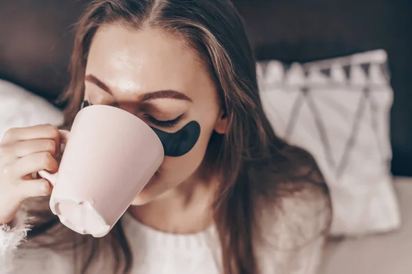 Trevlig ung tjej som vaknade tidigt på morgonen, drycker väldoftande kaffe, under de svarta fläckar mot ödem — Stockfoto