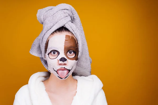 Ένα αστείο κορίτσι με μια ρόμπα terry λευκό και με μια πετσέτα στο κεφάλι της δείχνει τη γλώσσα, μια ενυδατική μάσκα με το πρόσωπο ενός σκύλου στο πρόσωπό της — Φωτογραφία Αρχείου