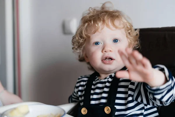 Regordete pequeño bebé con ojos azules sentado en la cocina comiendo una comida saludable —  Fotos de Stock