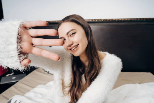 Piękna dziewczyna uśmiechający się z długimi włosami i w piżamie biały, siedząc na łóżku, zasłaniając twarz z jej strony z aparatu — Zdjęcie stockowe