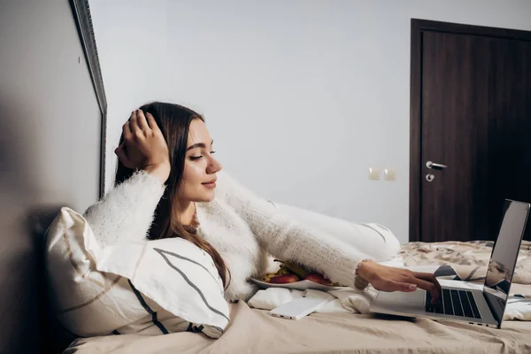 Junges schläfriges Mädchen, das spät in der Nacht auf dem Bett liegt und eine interessante Fernsehsendung auf ihrem Laptop anschaut — Stockfoto