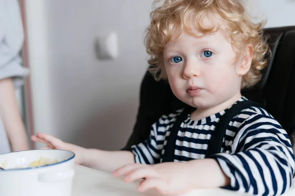 Маленький голубоглазый мальчик ест вкусную полезную кашу на кухне со своей матерью — стоковое фото