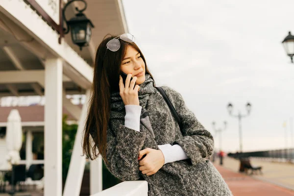 Une jolie fille aux cheveux bruns dans un manteau gris marchant dans les rues de la ville, profitant du temps nuageux et parlant au téléphone — Photo