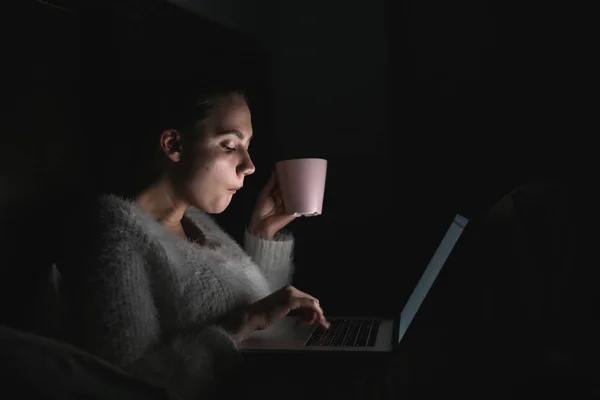 Gece geç saatte yatakta otururken, dizüstü bilgisayarda çalışan ve kahve içme kız serbest meslek — Stok fotoğraf