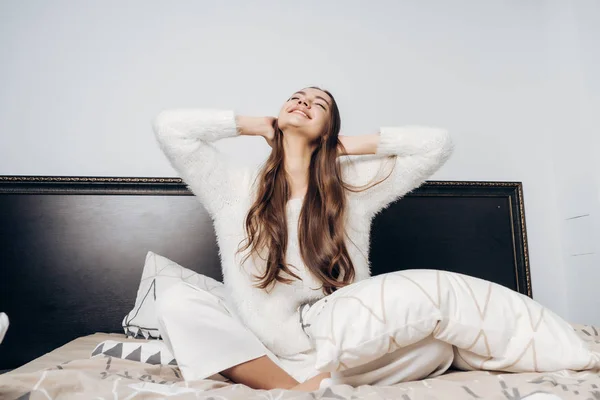 Glückliches junges Mädchen im weißen Pyjama sitzt im Bett, wacht früh morgens gut gelaunt auf — Stockfoto
