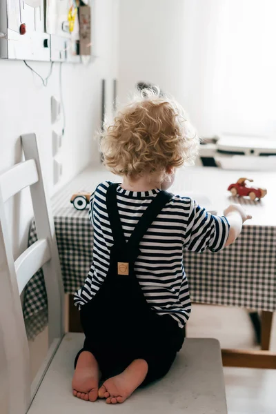 Niño rubio en ropa linda sentado en la silla cerca de stoloa, dibuja — Foto de Stock