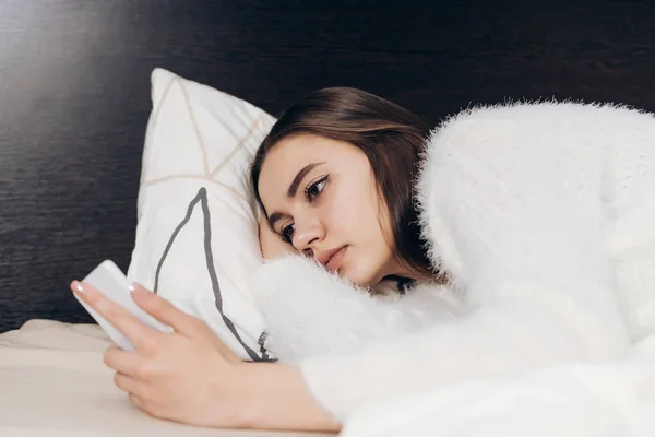 Sonnolenta ragazza in pigiama bianco si trova a letto la mattina presto, spegne l'allarme sul suo telefono — Foto Stock