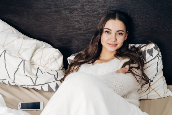 Hermosa joven en pijama blanco descansando en la cama después de un duro día de trabajo, sonriendo — Foto de Stock