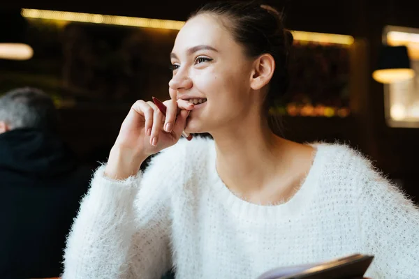 Милая молодая девушка в белом свитере сидит в кафе после работы, хорошее настроение, улыбается и смотрит в окно — стоковое фото