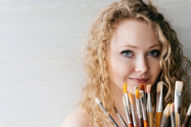 kıvırcık saçlı sevimli mavi gözlü kız çizim için fırça tutar, kamera ve gülümsüyor görünüyor