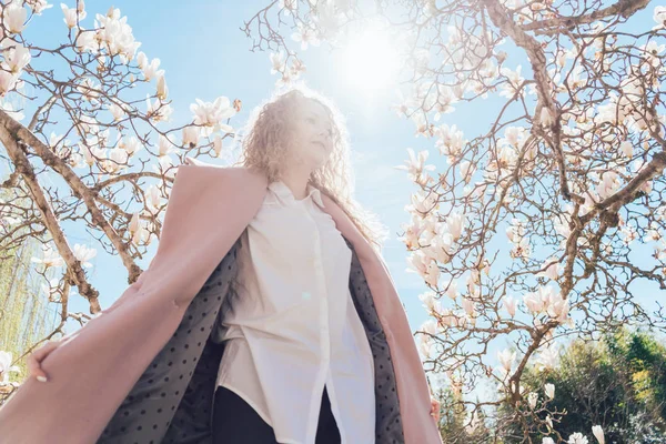 Elegante donna riccia in un cappotto rosa in posa contro un cielo blu e sole, accanto a una magnolia profumata — Foto Stock